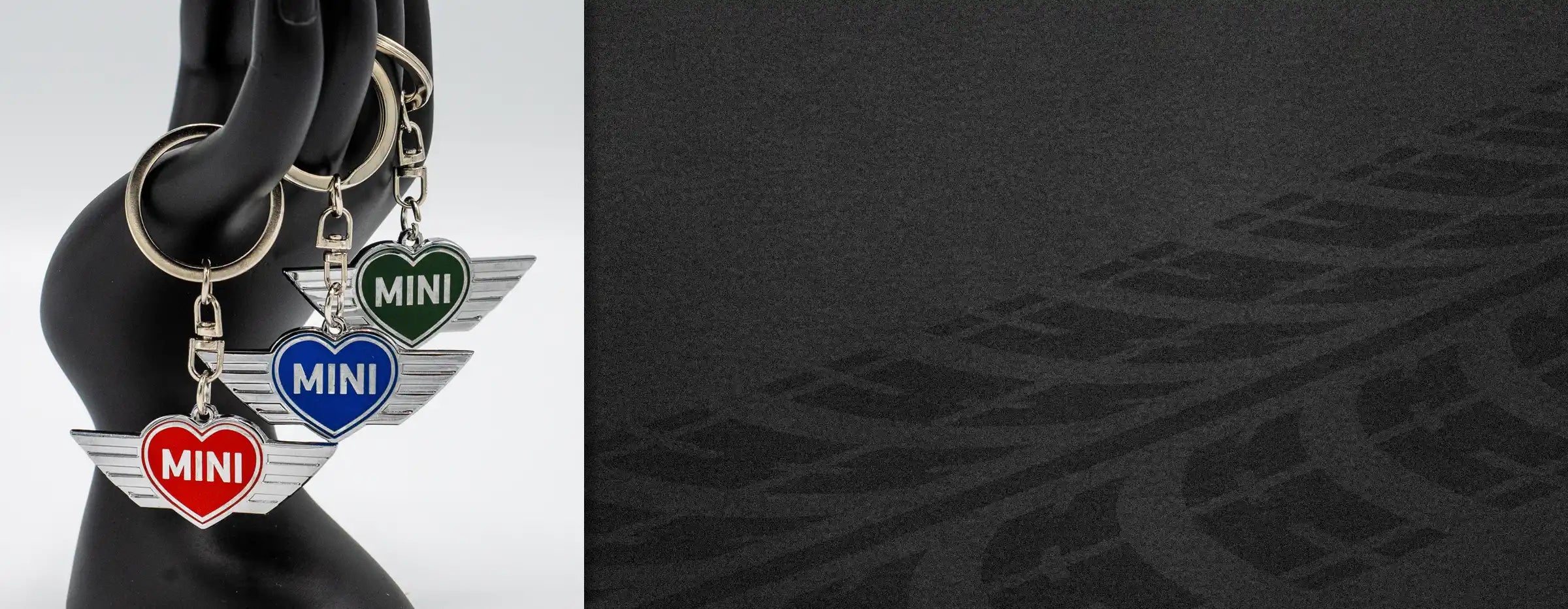 MINI heart wing keychains in chili red, electric blue, and British racing green displayed on a hand model against a dark textured background.